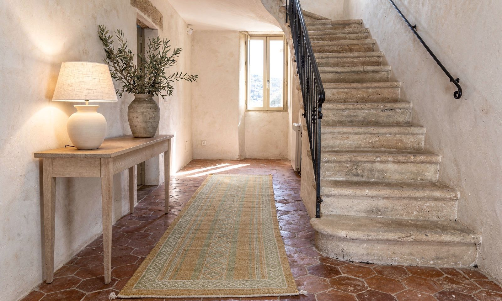 Bright hallway in a Grasse village house with a stone staircase, terracotta floor tiles and an ochre runner