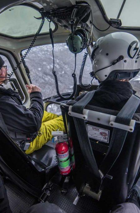 View from inside a helicopter cabin as the pilot and a helmeted passenger fly low over snow-covered mountain slopes.