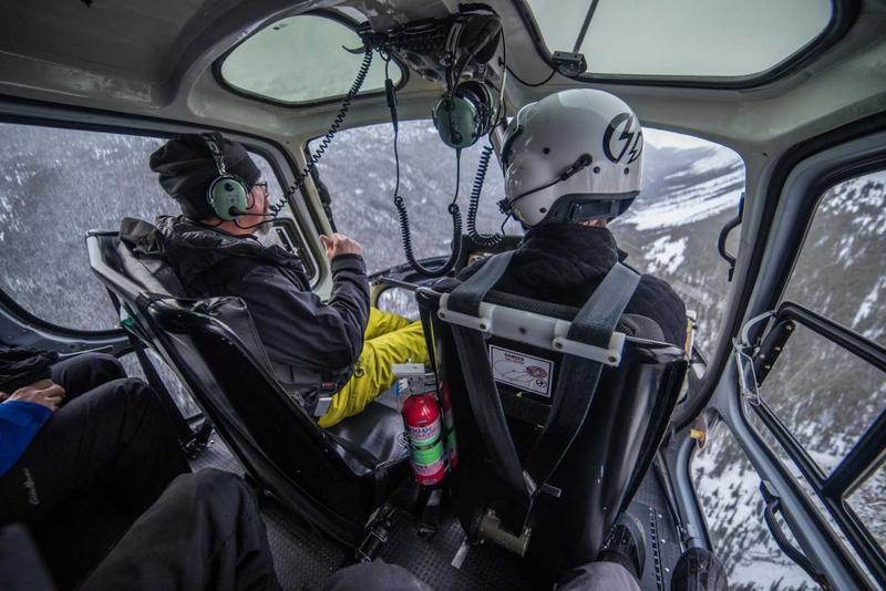 View from inside a helicopter cabin as the pilot and a helmeted passenger fly low over snow-covered mountain slopes.