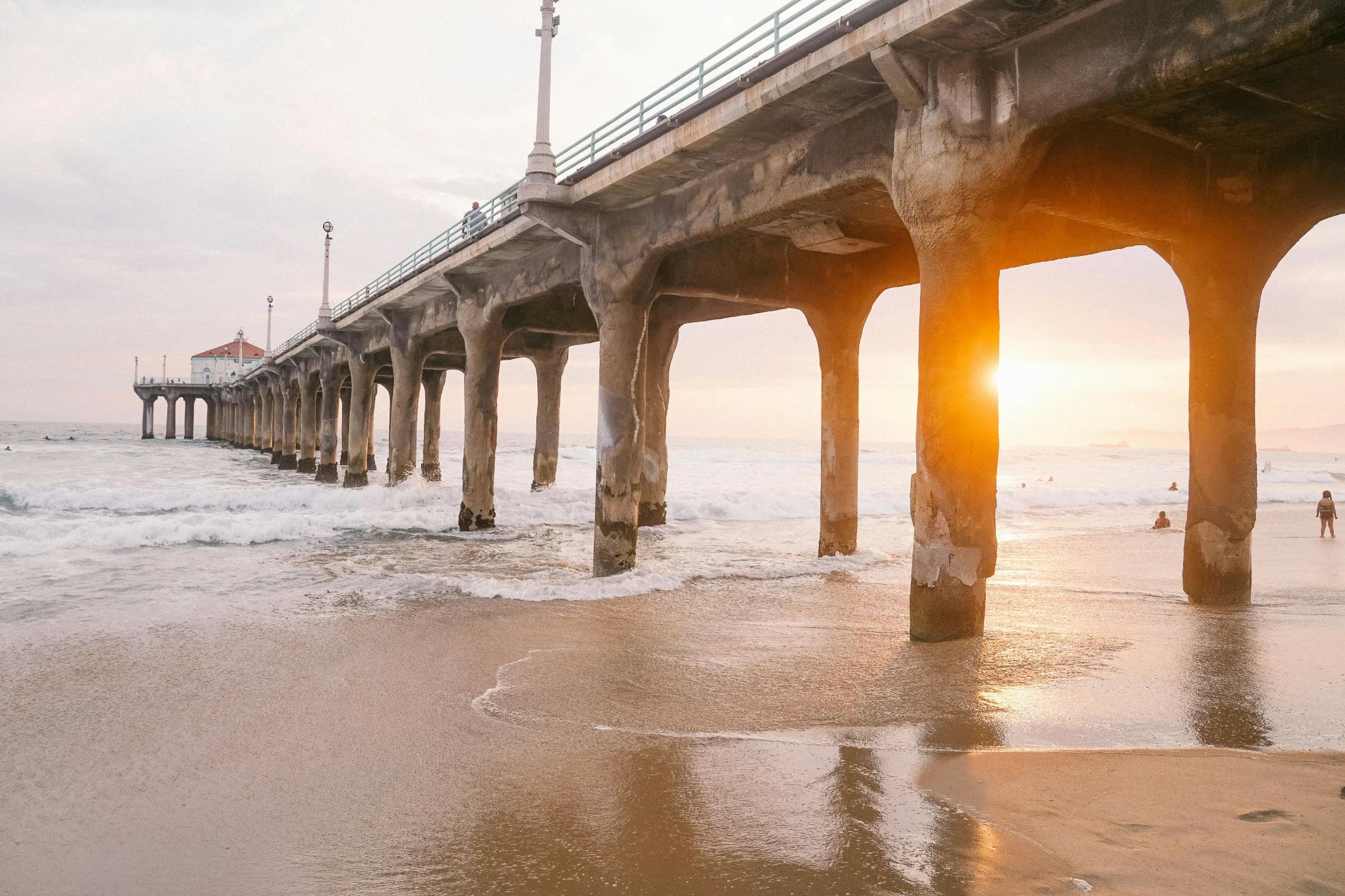 View along the underside of a pier at sunset, with waves washing beneath the concrete pilings and the sun glowing between them.
