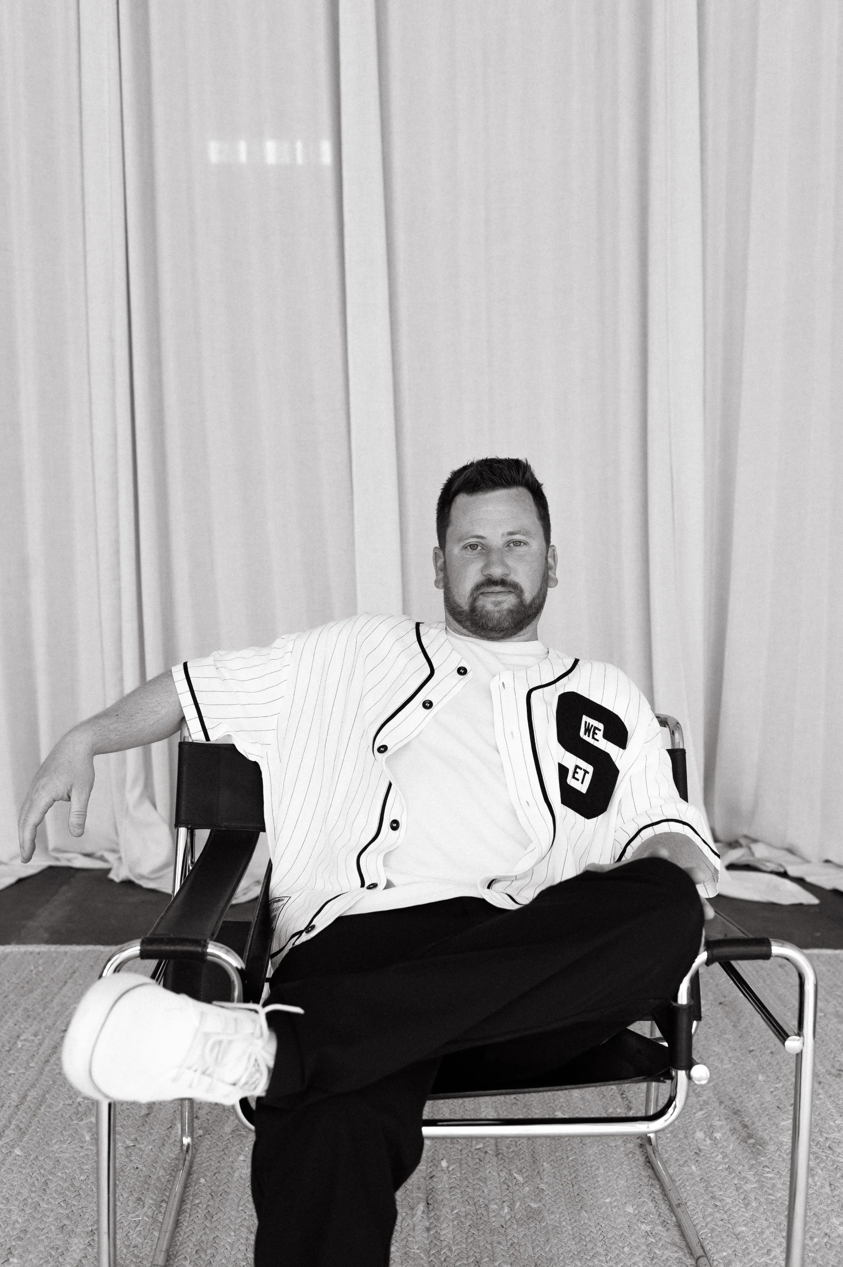 Black-and-white portrait of Alex Emberlin, Half + Half's production and video co-founder, seated in a director's chair against a curtained backdrop.