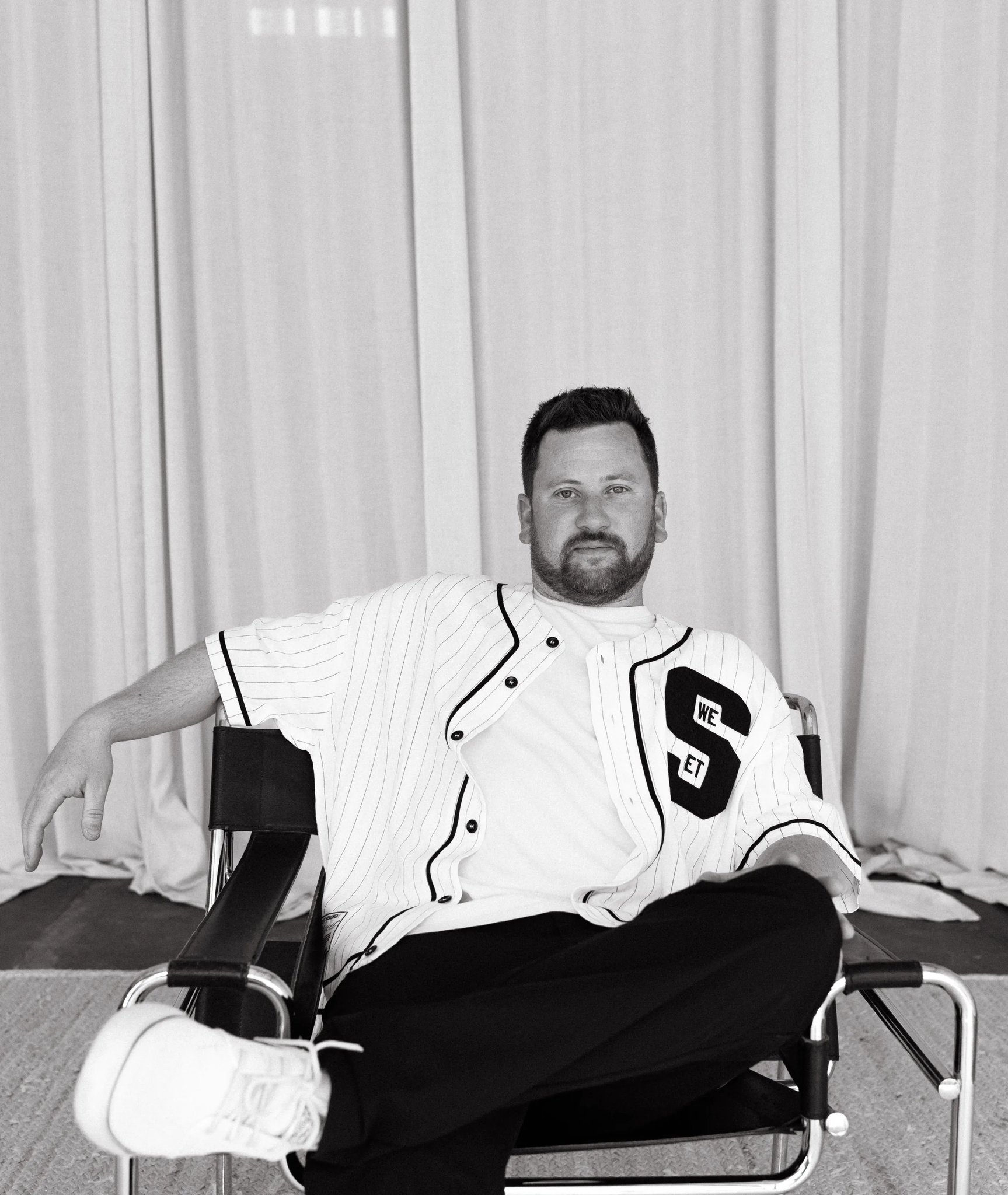 Black-and-white portrait of Alex Emberlin, Half + Half's production and video co-founder, seated in a director's chair against a curtained backdrop.