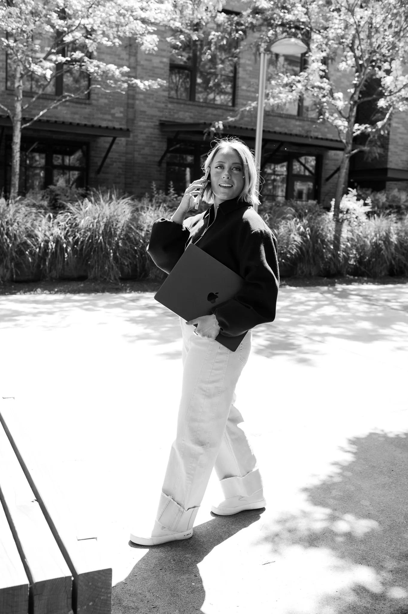 Black-and-white portrait of Olivia Krueger, Half + Half's branding and strategy co-founder, walking outdoors on a phone call while carrying a laptop.