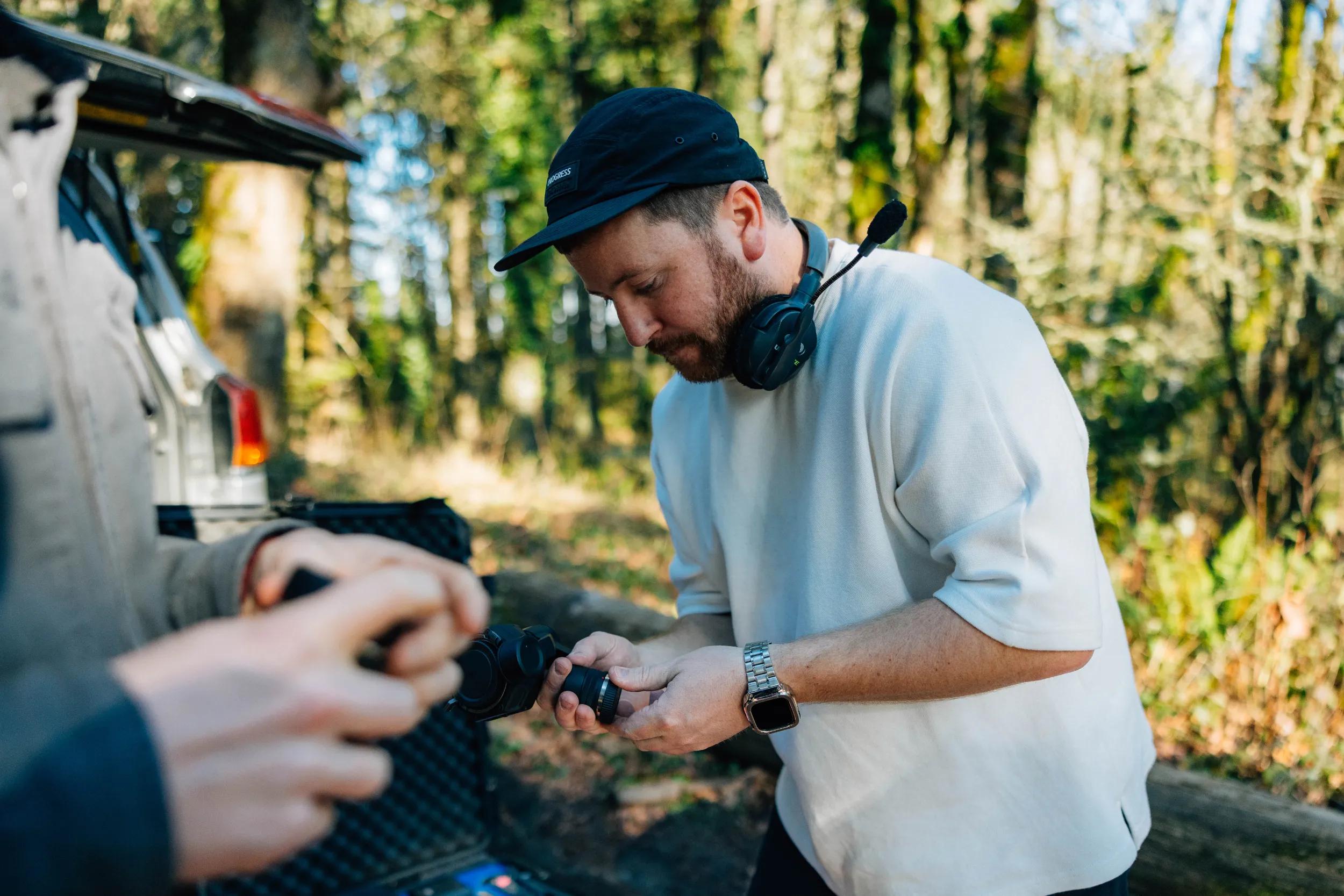 Man wearing a headset and cap changing a camera lens on a forest film set, illustrating the creative direction and photo/video service.
