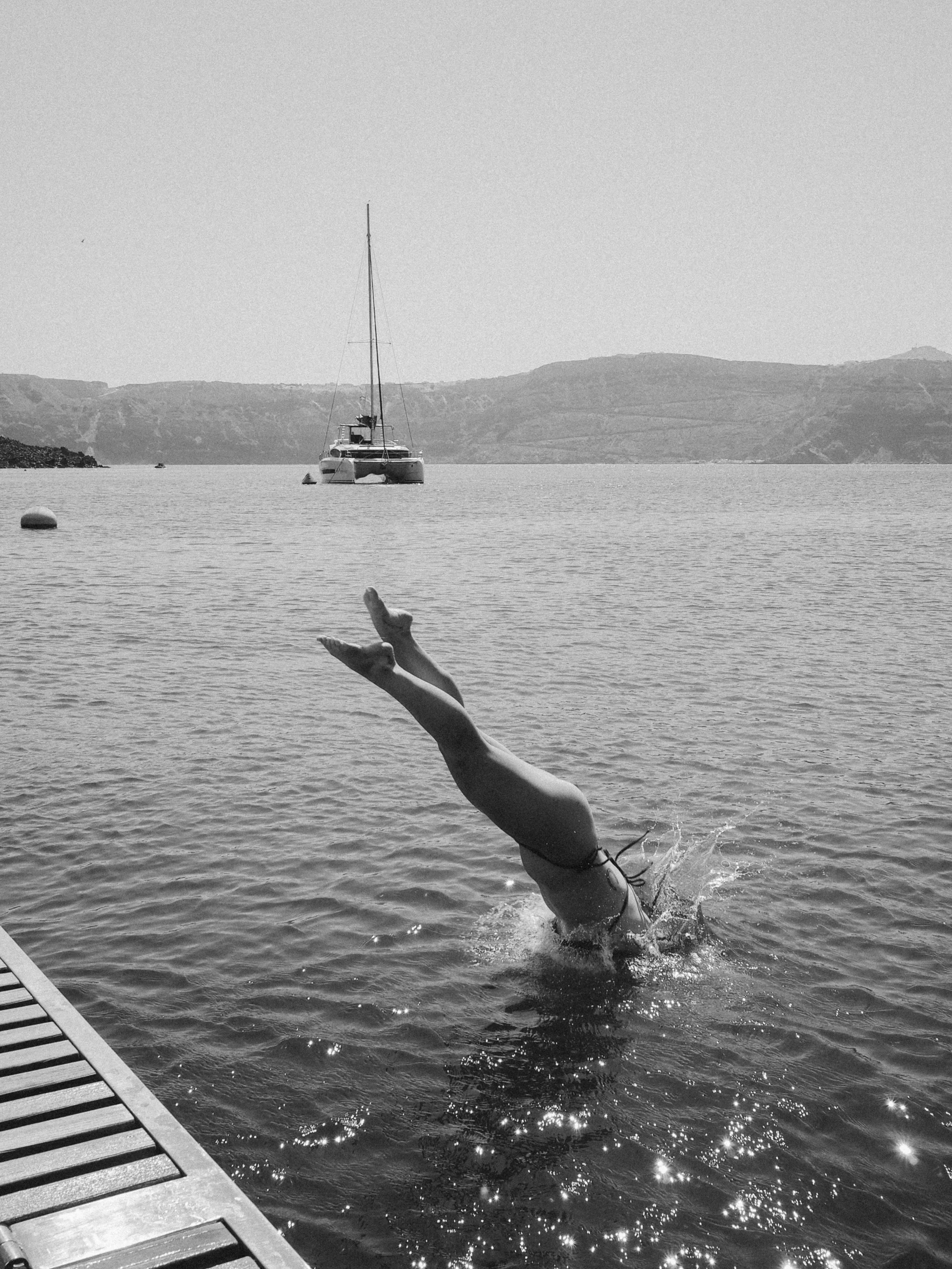 Black-and-white photo of a person diving off a dock into calm water, with a sailboat anchored in the bay behind them.