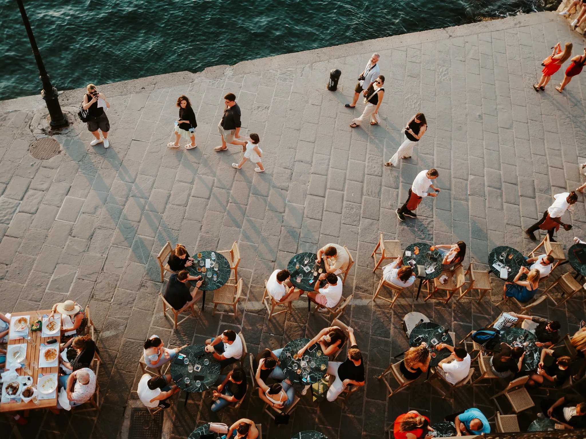 Aerial drone photograph looking straight down on a waterfront café terrace, with diners at tables and pedestrians walking along the stone promenade.