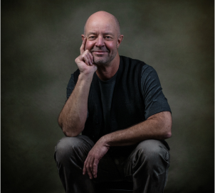 portrait of Richard smiling and sitting down leaning on his right knee with his elbow leading up to his head resting on his right hand while his left arm drapes comfortably across his lap. He's wearing jeans and a dark t-shirt.