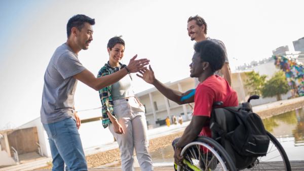 Amigos sorridentes ao ar livre; um deles, em cadeira de rodas, cumprimenta outro com um toque de mãos.