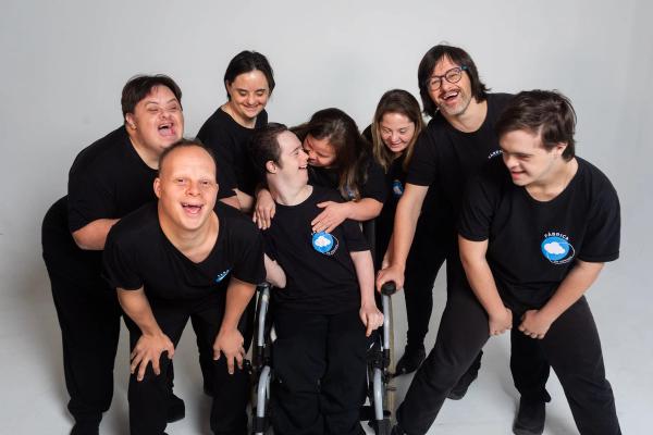 Elenco da Fábrica de Sonhos, com camisetas pretas do grupo, reunido e rindo num estúdio de fundo branco.
