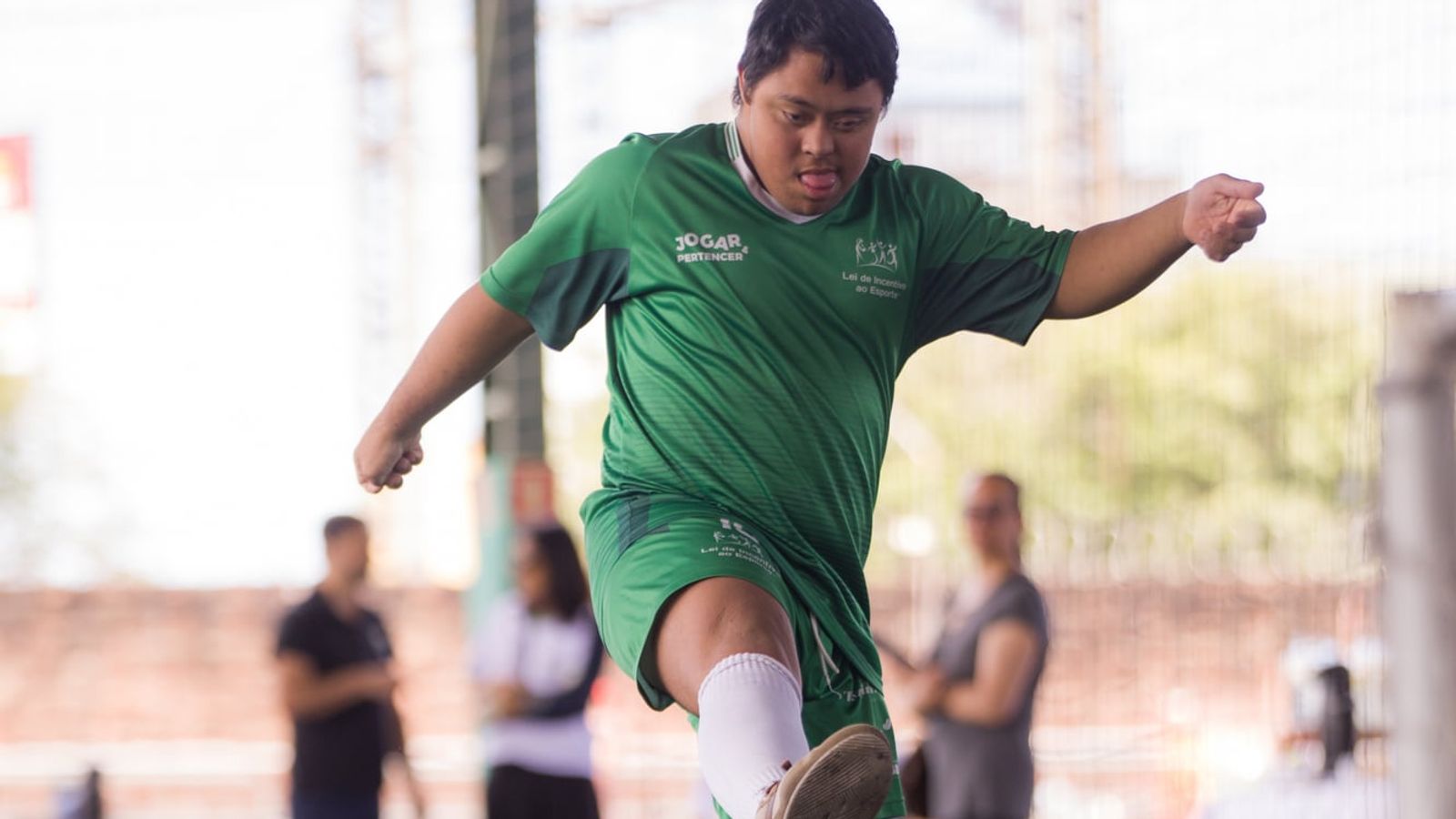 Jovem com síndrome de Down, de uniforme verde do projeto Jogar e Pertencer, chuta a bola num campo de grama sintética.