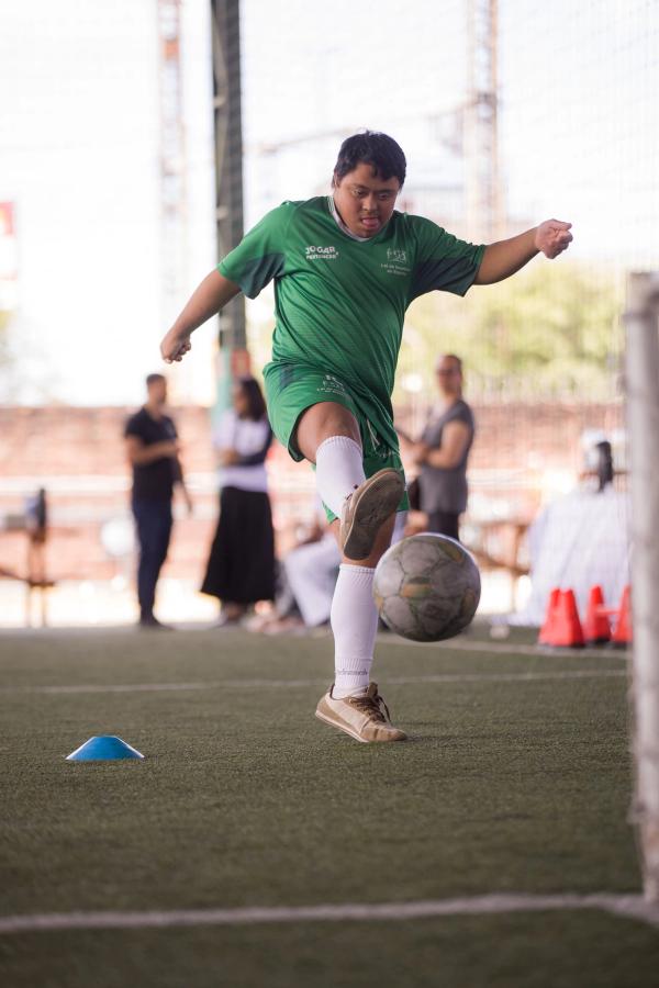 Jovem com síndrome de Down, de uniforme verde do projeto Jogar e Pertencer, chuta a bola num campo de grama sintética.