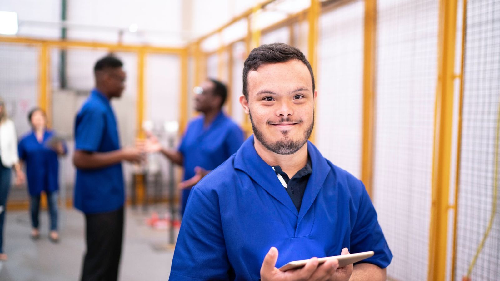 Jovem com síndrome de Down, de uniforme azul e tablet na mão, sorri num ambiente de trabalho; colegas conversam ao fundo.