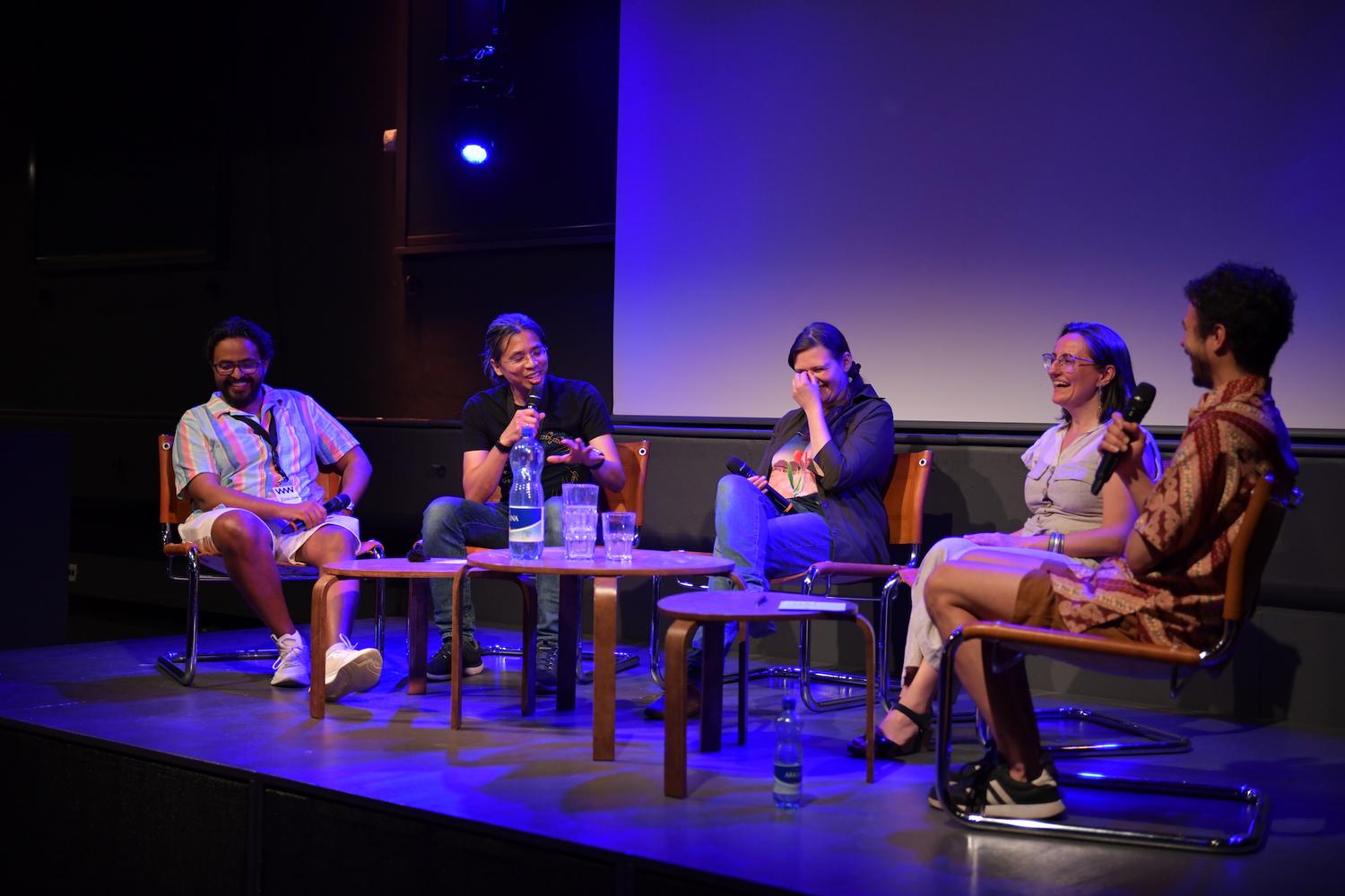 Eine Podiumsdiskussion mit fünf Personen, die auf einer Bühne sitzen und von blauem Licht beleuchtet werden. Sie sitzen in einem Halbkreis, haben Mikrofone in der Hand und sind in ein Gespräch und Lachen vertieft. Vor ihnen steht ein Tisch mit Wasserflaschen und Gläsern. Die Atmosphäre ist informell und entspannt, die Teilnehmer teilen einen lockeren Moment. Der Hintergrund ist dunkel, und der Fokus liegt auf den Teilnehmern und der Unterhaltung.
