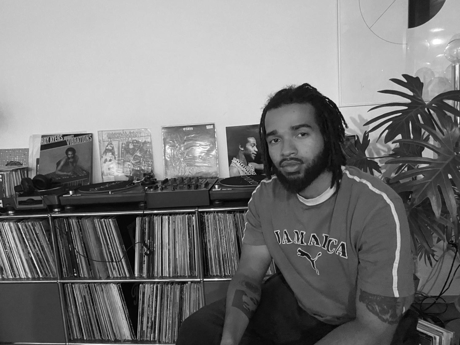Eine schwarzweiss Fotografie. In der rechten Bildhälfte sitzt eine Schwarze Person mit Bart und mittellangen Dreadlocks. Dahinter ein Regal mit Musikplatten. 