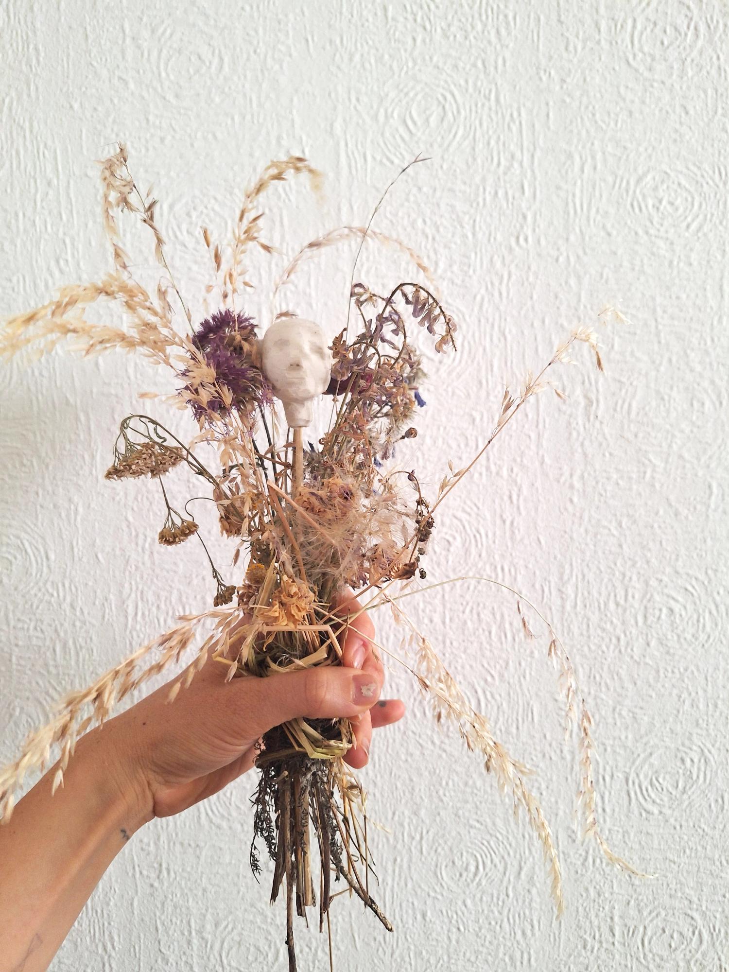 Eine Hand hält einen Strauss aus getrockneten Gräsern und Blumen, in dem eine kleine menschliche Figur mit Kopf aus Ton oder Pappmaché steckt. Im Hintergrund eine weisse Raufasertapete. 