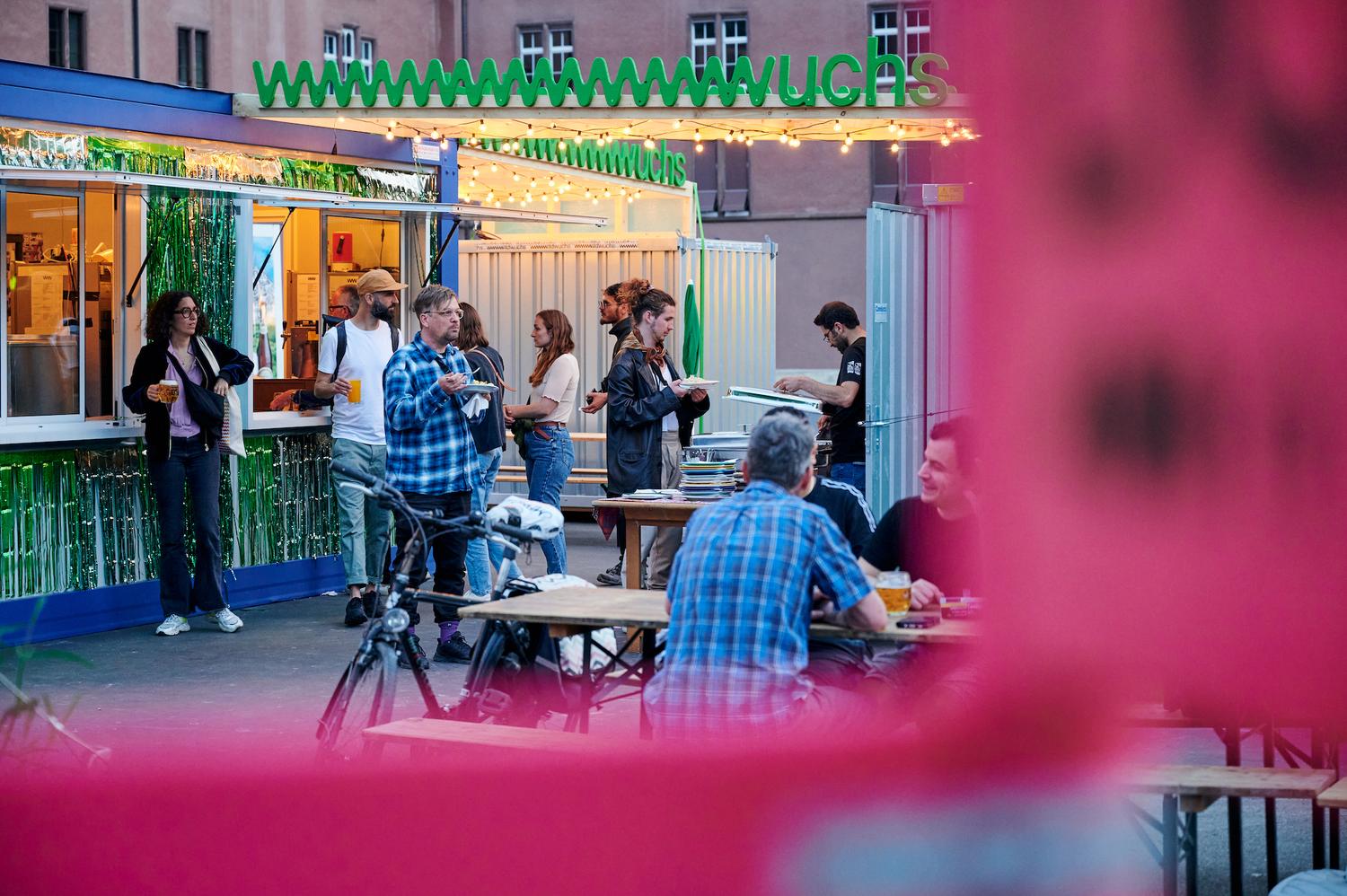 Das Bild zeigt das Fewstivalzentrum von Wildwuchs. Zu sehen ist ein mit grünem Lametta behangener Bar-Container, sowie einige Bänke und sitzende und stehende Menschen.