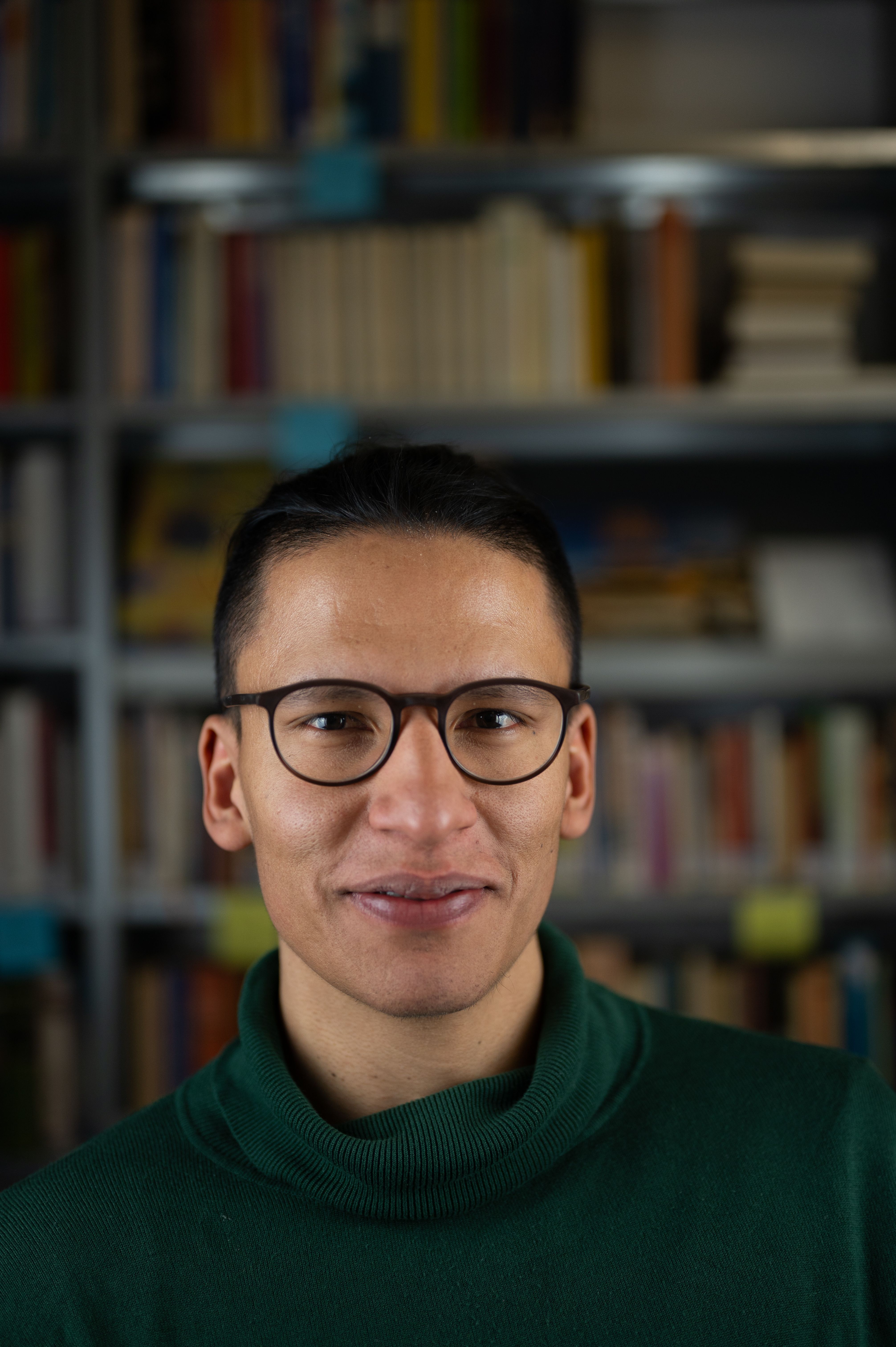 Porträt einer männlich gelesenen Person mit Brille und dunklen Haaren. Er lächelt in die Kamera. 