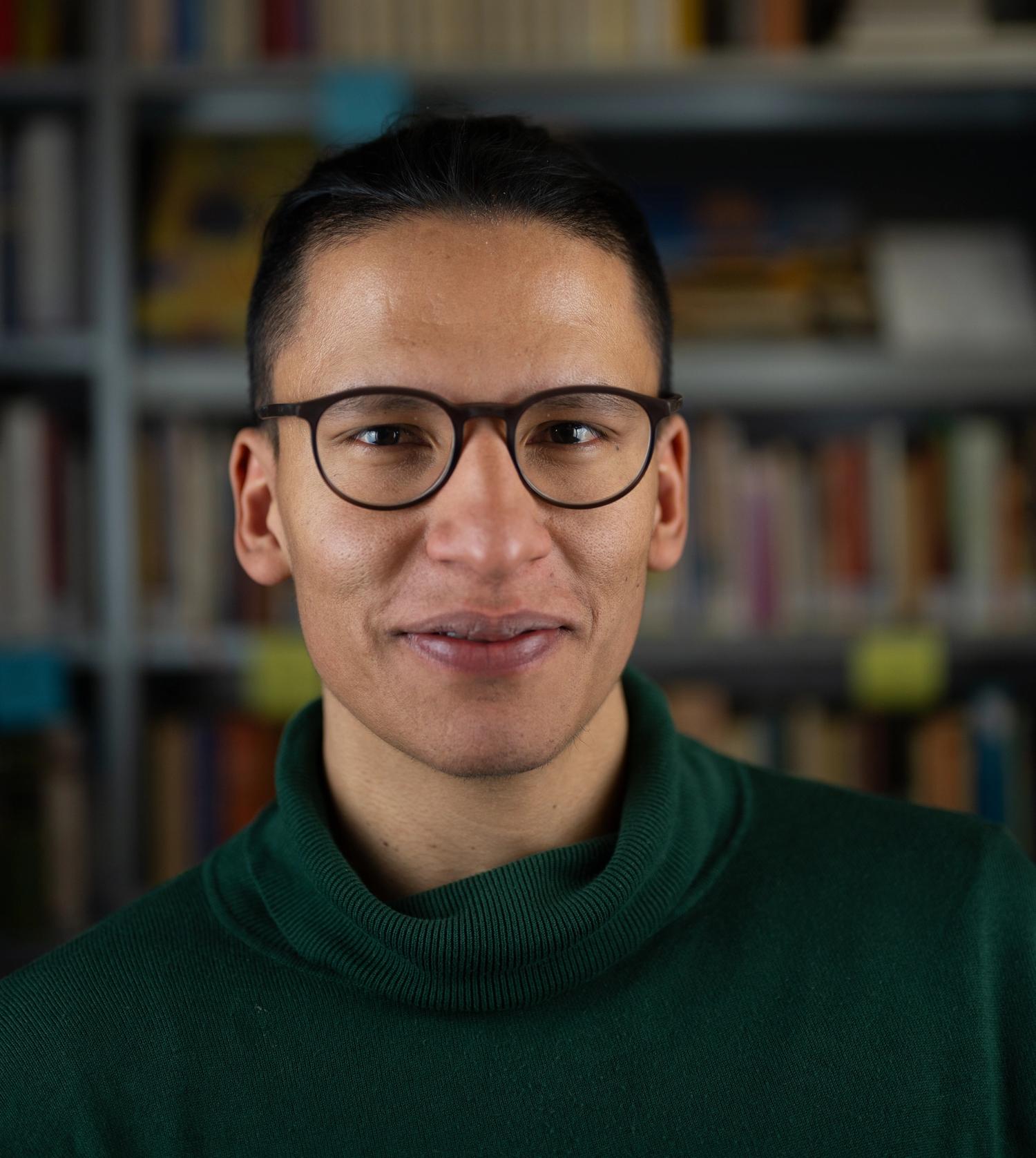 Porträt einer männlich gelesenen Person mit Brille und dunklen Haaren. Er lächelt in die Kamera.