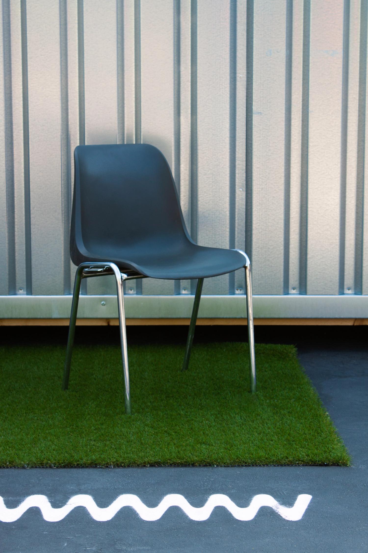 Auf dem Foto ist ein leerer Stuhl zu sehen. Der Stuhl ist grau und steht auf einem Rasenteppich und vor einer silbernen Wand aus Aluminium. Vor dem Rasenteppich ist das Wildwuchslogo auf dem Asphaltboden gesprayt. Das Foto wurde am Wildwuchs Festival 2023 aufgenommen. 