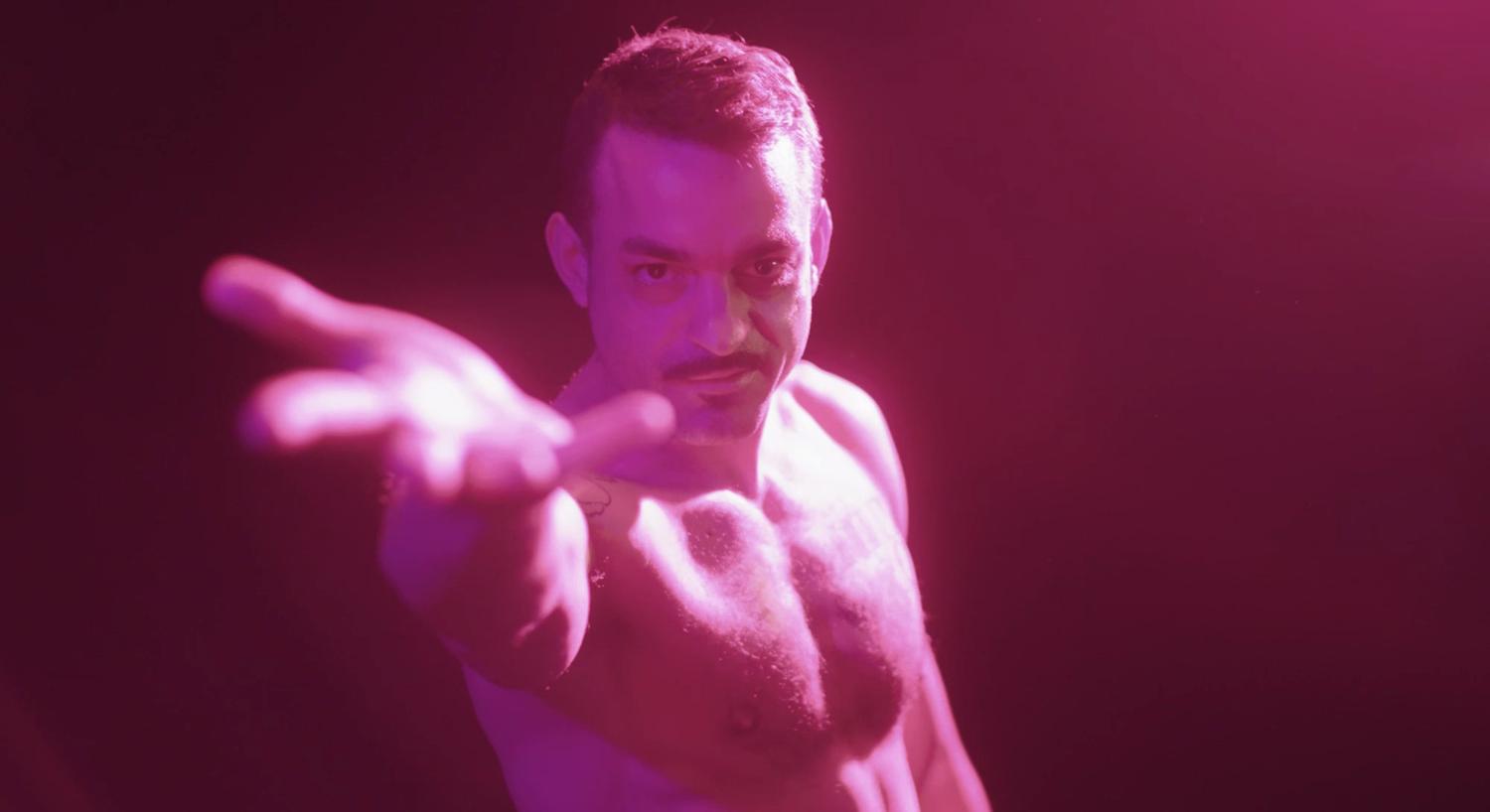 Ein dramatisch beleuchtetes Foto mit rosa Lichtbeleuchtung eines Mannes ohne Hemd, dessen Hand mit der Handfläche nach oben in Richtung Kamera reicht