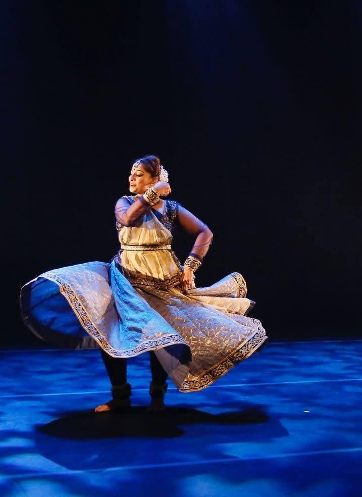 Tanzende Person auf einer Bühne mit dunklem Hintergrund und blauem Bodenlicht. Die Person trägt ein traditionelles, reich verziertes Kleid in Blau und Gold, das sich beim Drehen weit auffächert. Die Pose ist elegant, mit einem Arm vor dem Gesicht und dem anderen zur Seite ausgestreckt, begleitet von ausdrucksstarker Mimik.
