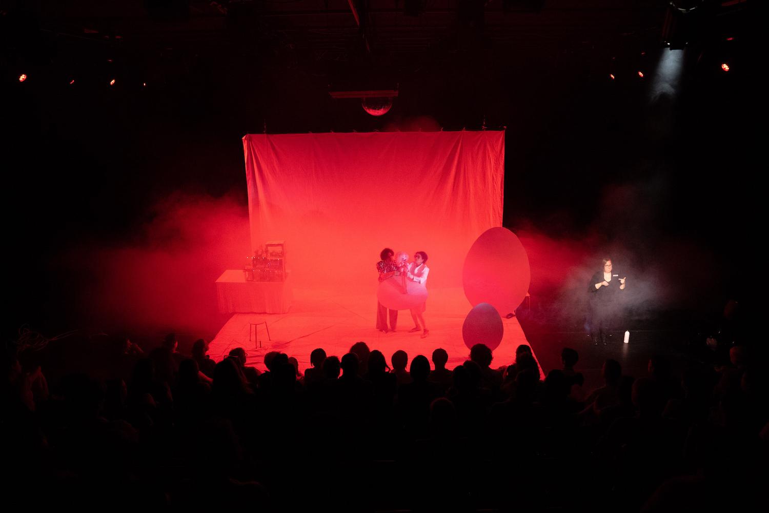 Eine Theateraufführung in einem dunklen Auditorium, das durch dramatisches rotes Licht beleuchtet wird. Zwei Darsteller stehen im Zentrum der Bühne und halten einen durchscheinenden Gegenstand zwischen sich, im Hintergrund ein roter Vorhang. Auf der Bühne befinden sich runde rote Requisiten und ein Tisch mit Gegenständen links im Bild. Das Publikum ist als Silhouette zu erkennen, das die Szene aufmerksam verfolgt. Rechts im Bild führt eine Person Gebärdensprache auf. Rauch oder Nebel verstärkt die Atmosphäre und erzeugt eine surreale, intensive Stimmung.