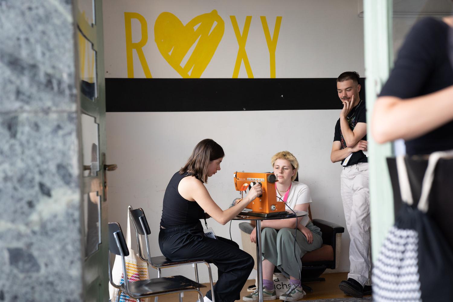 Eine Szene in einem Raum, in dem drei Personen um eine orangefarbene Nähmaschine versammelt sind. Eine Person sitzt an der Nähmaschine, während eine andere Person gegenüber sitzt und aufmerksam zuschaut. Eine dritte Person steht in der Nähe und beobachtet die Aktivität interessiert. Im Hintergrund ist eine Wand mit dem Wort “ROXY” in fetten gelben Buchstaben und einem Herzsymbol, das über einem schwarzen horizontalen Streifen gemalt ist.