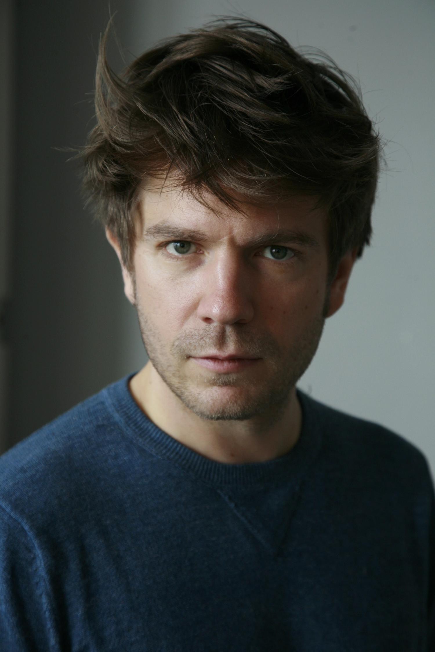 Porträtaufnahme einer Person mit kurzem, zerzaustem braunem Haar, leichtem Bartschatten und ernstem Gesichtsausdruck. Die Person trägt einen dunkelblauen Pullover und blickt direkt in die Kamera. Der Hintergrund ist neutral und unscharf.