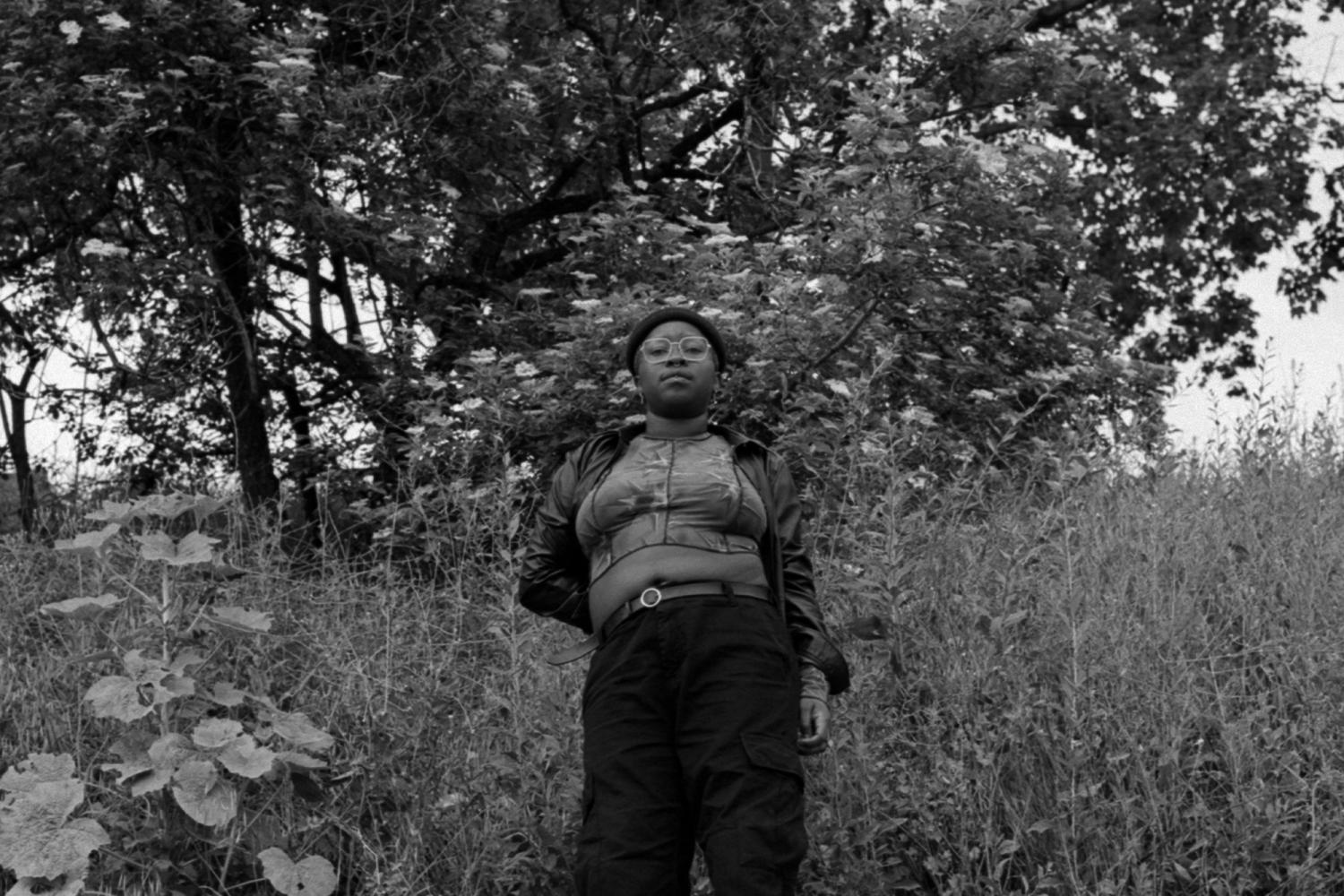 Eine Schwarze Person steht vor einem Baum und einer Wiese und blickt in die Kamera, leicht von unten fotografiert und ab Kniehöhe. 