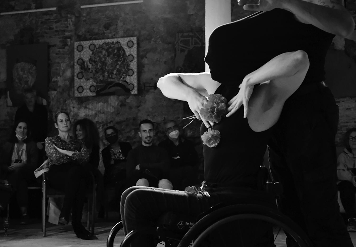 Eine Schwarz-Weiss-Fotografie zeigt eine von der Seite abgebildete tanzende Person, die im Rollstuhl sitzt. Die Hände sind zur Brust gewendet und halten Blumen. Das Gesicht ist vollständig von einem schwarzen Stoff bedeckt. Im Hintergrund ist ein Publikum zu erkennen. 