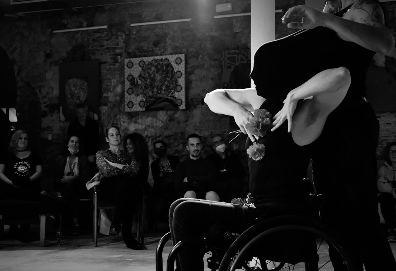 Eine Schwarz-Weiss-Fotografie zeigt eine von der Seite abgebildete tanzende Person, die im Rollstuhl sitzt. Die Hände sind zur Brust gewendet und halten Blumen. Das Gesicht ist vollständig von einem schwarzen Stoff bedeckt. Im Hintergrund ist ein Publikum zu erkennen. 