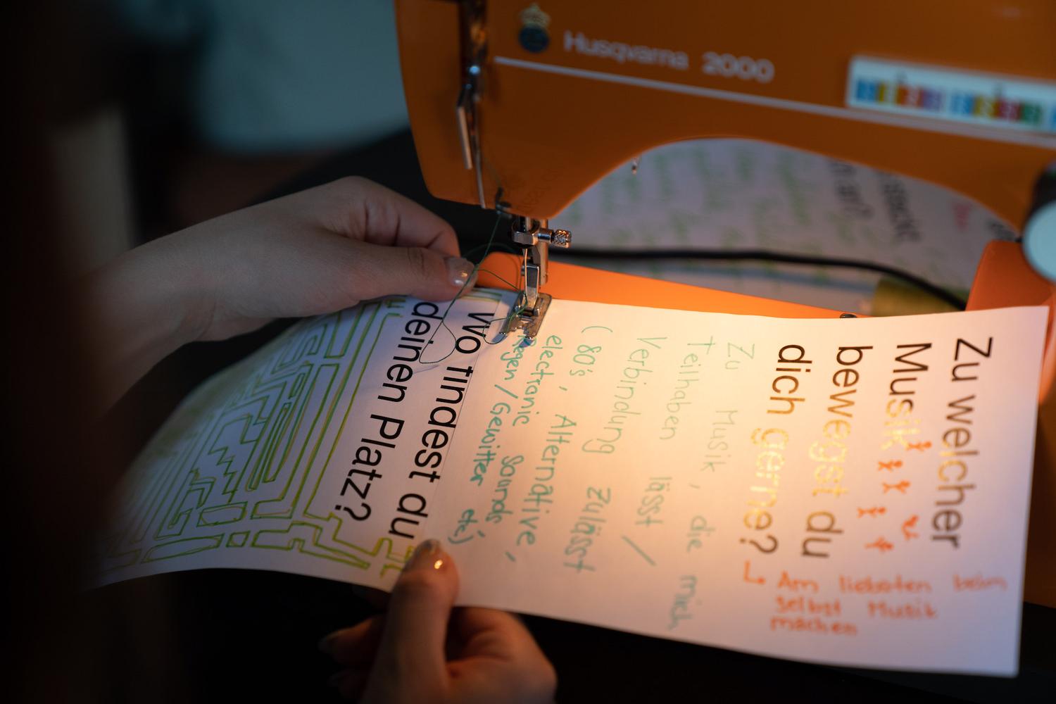 Nahaufnahme von Händen, die ein Blatt Papier durch eine orangefarbene Nähmaschine führen. Auf dem Papier sind handgeschriebene und gedruckte Texte in deutscher Sprache zu sehen, darunter “Zu welcher Musik bewegst du dich gerne?” und “Wo findest du deinen Platz?”. Die Nähmaschine näht das Papier mit grünem Faden.