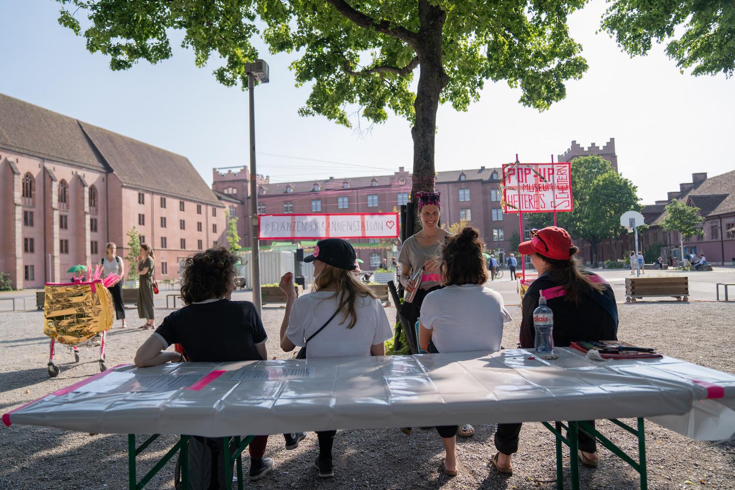 Eine Außenszene am Tag, bei der mehrere Personen an einem Tisch mit Plastikabdeckung unter einem schattenspendenden Baum sitzen. Sie blicken auf eine Frau, die vor ihnen steht, lächelt und Gegenstände in den Händen hält. Hinter ihr sind leuchtend pinke Schilder mit handgeschriebenen Texten zu sehen, darunter “Pop-Up Museum” und “Pflanzenstation”. Im Hintergrund sind rote Backsteingebäude und weitere Teilnehmer bei Aktivitäten zu erkennen, darunter ein mit goldener Folie bedeckter Einkaufswagen, der mit pinken Federn dekoriert ist.