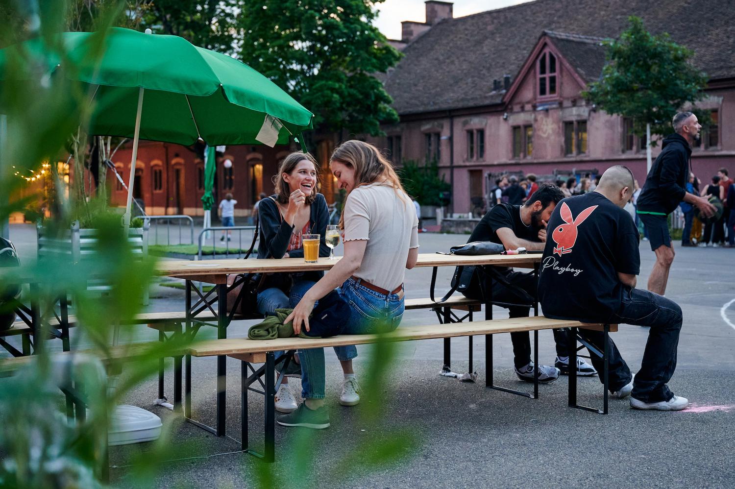 Eine Outdoor-Szene bei Dämmerung. Zwei Frauen sitzen an einer Holzbank im Picknickstil unter einem grünen Sonnenschirm, lachen und unterhalten sich bei Getränken. In der Nähe sitzen zwei Männer an derselben Bank, einer trägt ein schwarzes T-Shirt mit einem großen roten Playboy-Logo. Im Hintergrund ist ein Hofbereich mit Bäumen und einer Gruppe von Menschen vor einem roten Gebäude zu sehen, das sanfte Abendlicht schafft eine entspannte Atmosphäre.