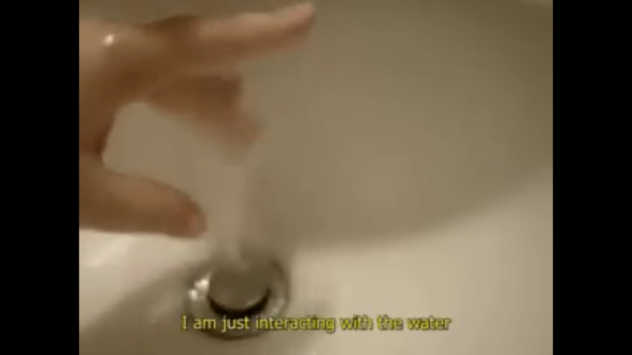 Unscharfes Bild einer Hand, die unter fliessendem Wasser in einem Waschbecken bewegt wird. Im unteren Bereich des Bildes ist ein gelber Untertitel auf Englisch eingeblendet: "I am just interacting with the water".