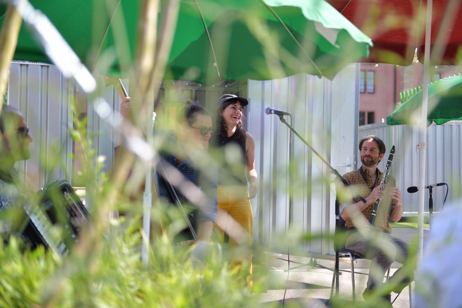 Ein Open-Air-Musikauftritt mit Musikern, die unter grünen Sonnenschirmen sitzen. In der Mitte steht eine Frau und lacht in der Nähe eines Mikrofons, während ein Mann rechts von ihr Klarinette spielt. Ein weiterer Musiker mit einem Akkordeon ist links teilweise sichtbar. Die Szene ist von Grünpflanzen im Vordergrund umrahmt und vermittelt eine entspannte, natürliche Atmosphäre. Im Hintergrund ist eine Metallwand zu sehen, und das Sonnenlicht sorgt für eine warme, helle Stimmung.