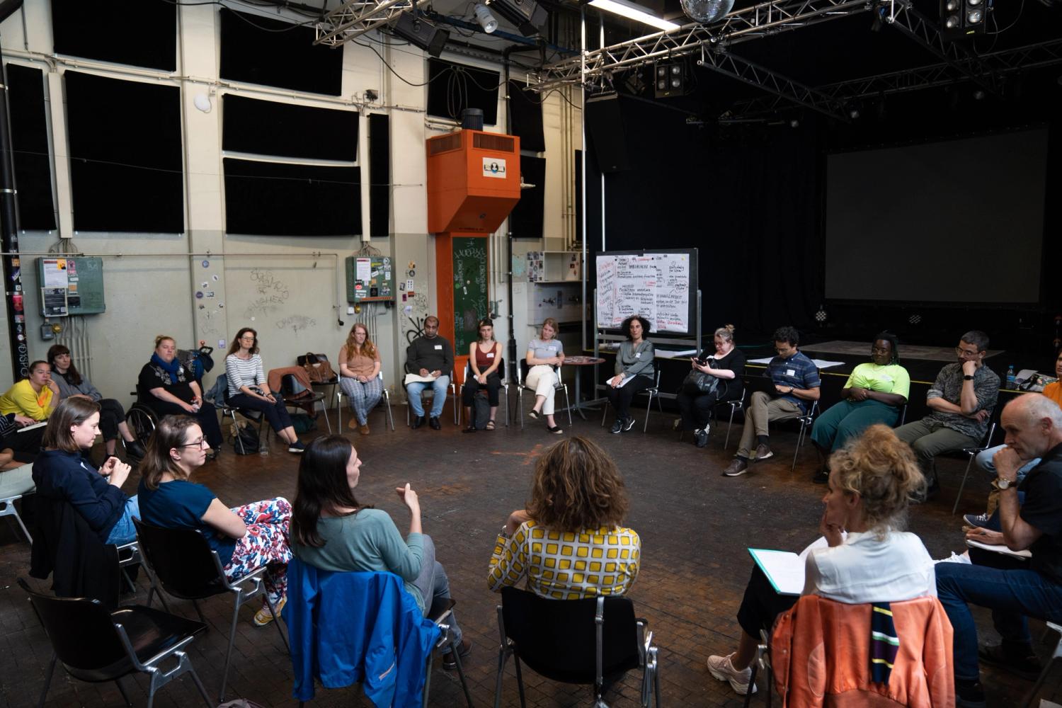 Auf dem Foto sitzen Menschen in einem Stuhlkreis und diskutieren miteinander. Es sind 23 Personen sichtbar. Hinter der Moderatorin steht eine Tafel an der vollgeschriebene Plipchart Blätter befestigt sind. 