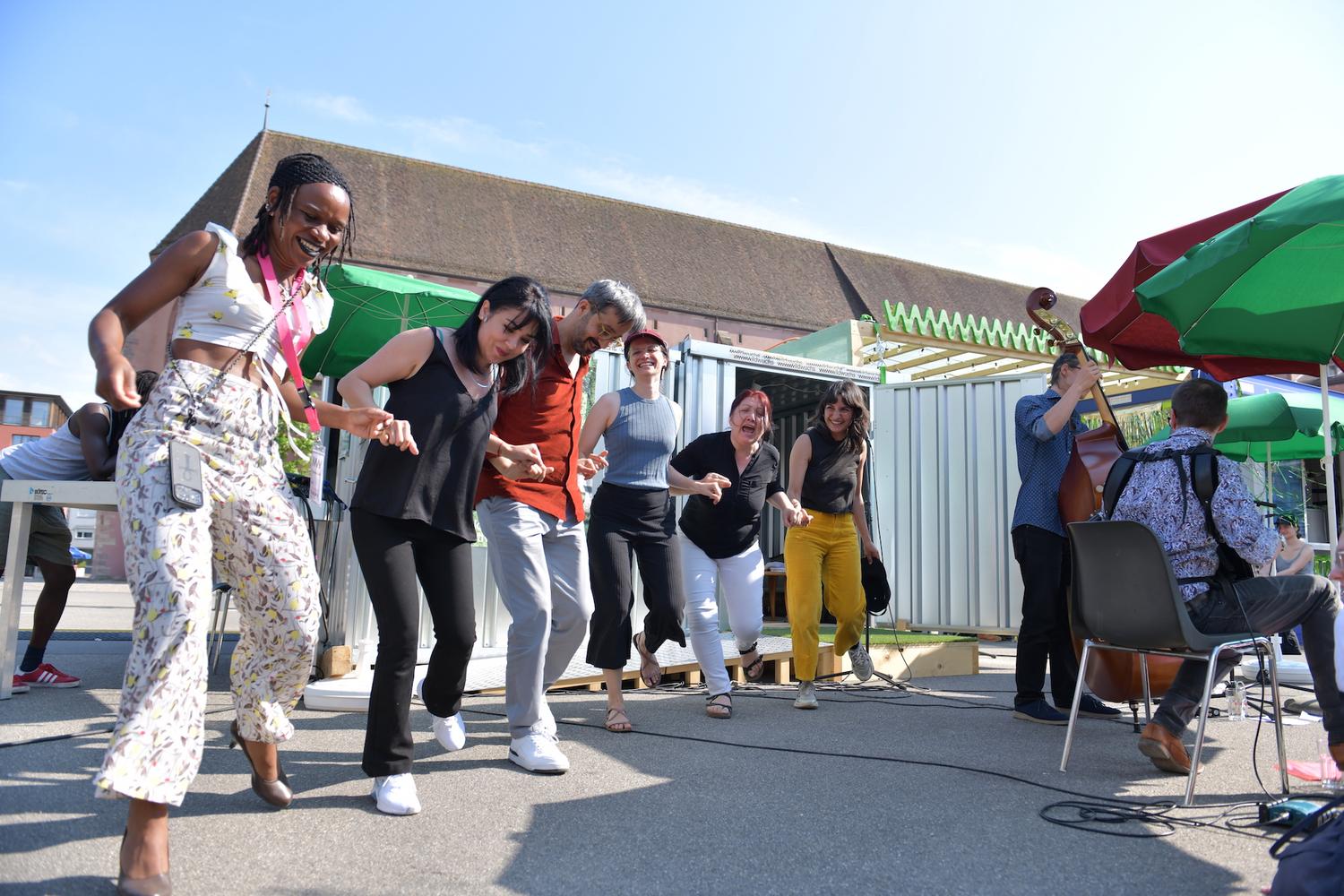 Eine Gruppe von Menschen tanzt im Freien und hält sich dabei an den Händen. Sie scheinen sich zu amüsieren, lachen und lächeln, während sie gemeinsam tanzen. Die Szene spielt sich im strahlenden Sonnenlicht mit klarem Himmel ab. Im Hintergrund spielt ein Musiker einen Kontrabass, während ein anderer Musiker in der Nähe sitzt. Grüne und rote Sonnenschirme spenden Schatten, und im Hintergrund ist eine Metallkonstruktion zu sehen. Die Stimmung ist lebhaft und festlich.