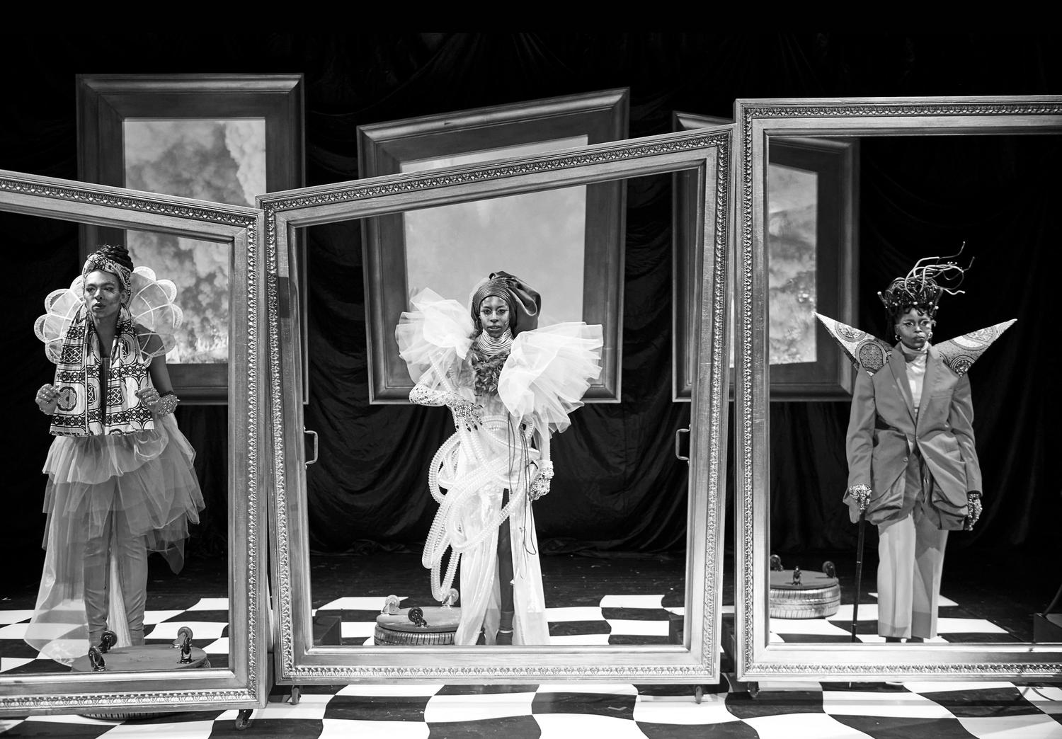 Eine Schwarz-Weiß-Fotografie zeigt eine szenische Inszenierung. Drei Personen in üppigen, prachtvollen Kostümen stehen Seite an Seite auf einer Bühne, lebensgroße Gemälderahmen umranden sie.