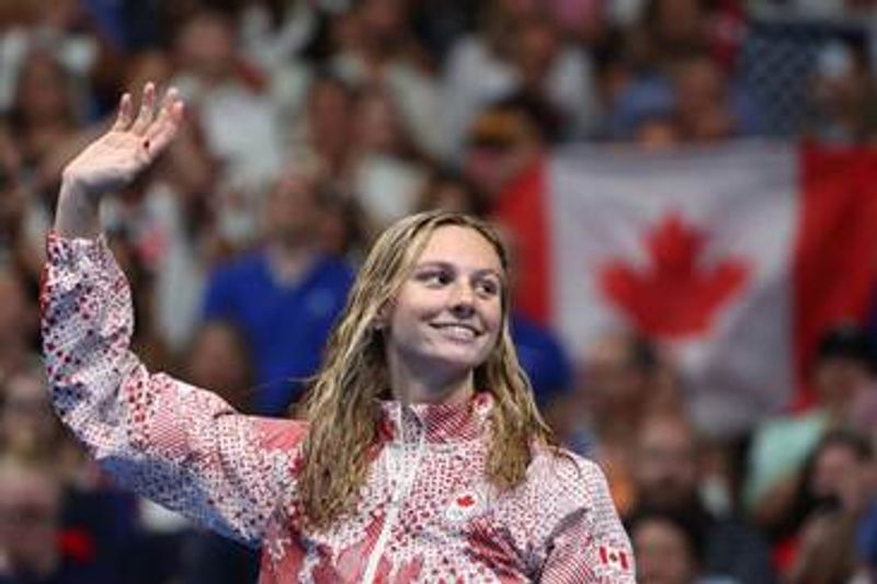 Summer McIntosh, nageuse triple championne olympique, lève la main, un drapeau du canada en fond.