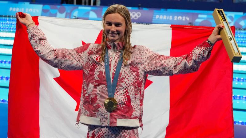 Summer McIntosh, nageuse triple championne olympique, porte une médaille d'or, un drapeau du canada derrière elle.