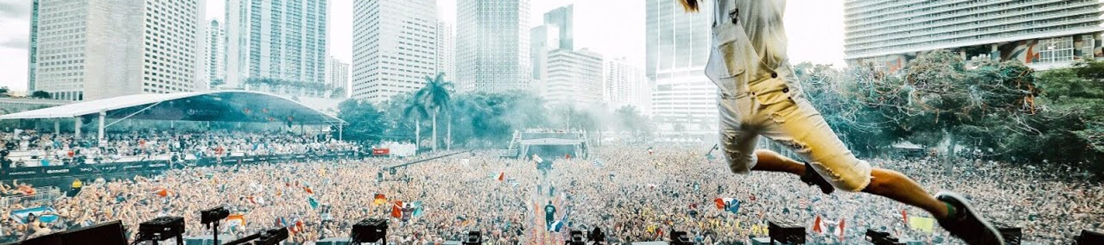 Steve Aoki Leaping into the DJ Book The image captures an energetic scene at a large outdoor music festival, likely Ultra Music Festival in Miami, given the iconic downtown skyline in the background.
