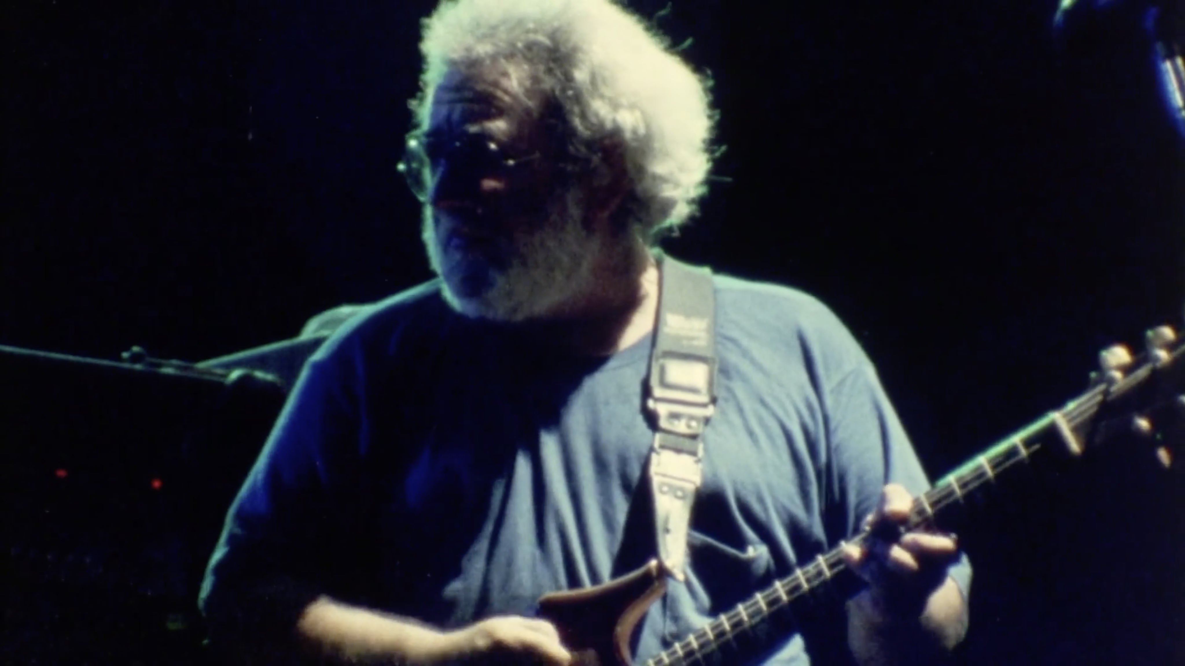 A medium close-up, dimly lit photograph of Jerry Garcia performing on stage. He wears his signature glasses and has a thick, bushy white beard. He is focused on playing his electric guitar, with his fingers positioned on the fretboard. The lighting is dark with a cool blue cast, highlighting his face and the instrument against a dark background.