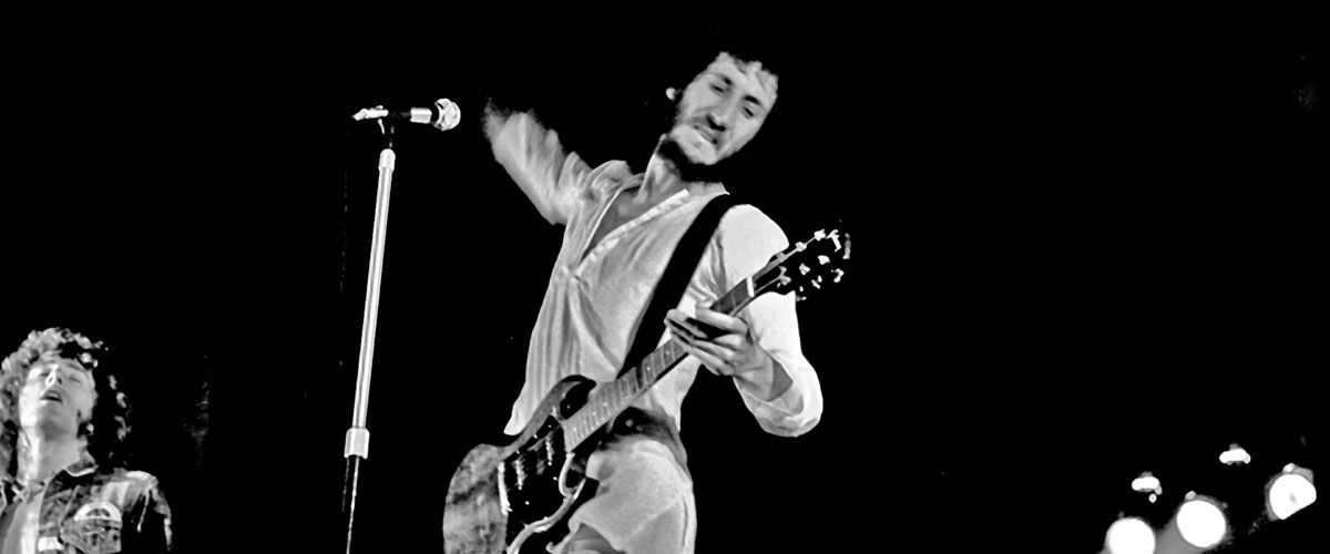 A high-contrast black and white photograph of Pete Townshend, guitarist for The Who, performing live on stage.  Townshend is captured in a dynamic, mid-action pose, leaning back with a focused expression. He is wearing a white button-down shirt and holding a dark electric guitar, likely a Gibson SG. His right arm is raised high behind him, caught in the middle of his signature "windmill" strumming motion.