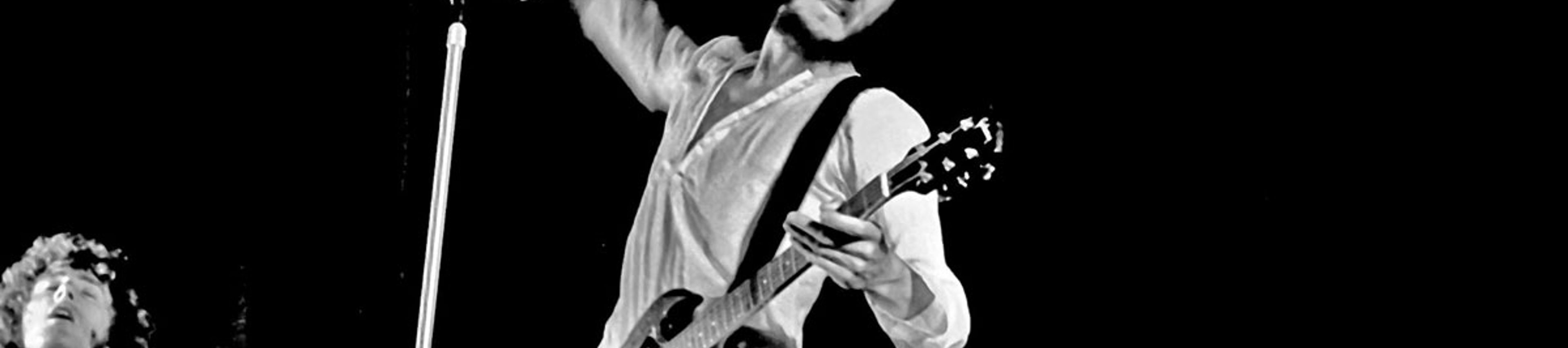 A high-contrast black and white photograph of Pete Townshend, guitarist for The Who, performing live on stage. Townshend is captured in a dynamic, mid-action pose, leaning back with a focused expression. He is wearing a white button-down shirt and holding a dark electric guitar, likely a Gibson SG. His right arm is raised high behind him, caught in the middle of his signature "windmill" strumming motion.