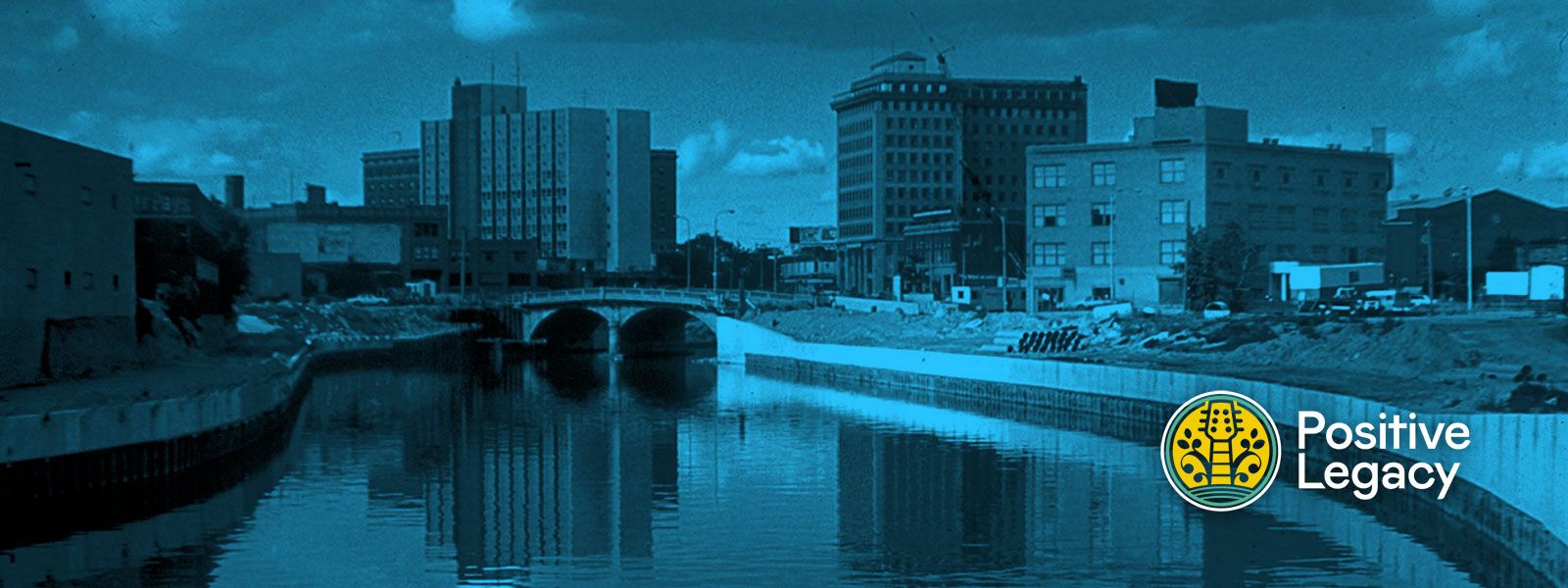 A wide, panoramic landscape photo of a city canal or riverfront, treated with a moody teal-blue tint.  The scene features a calm body of water in the foreground reflecting the sky and buildings. A concrete arched bridge crosses the water in the mid-ground. Along the banks, there are several multi-story urban buildings, some appearing older and industrial, set against a sky with soft, scattered clouds.