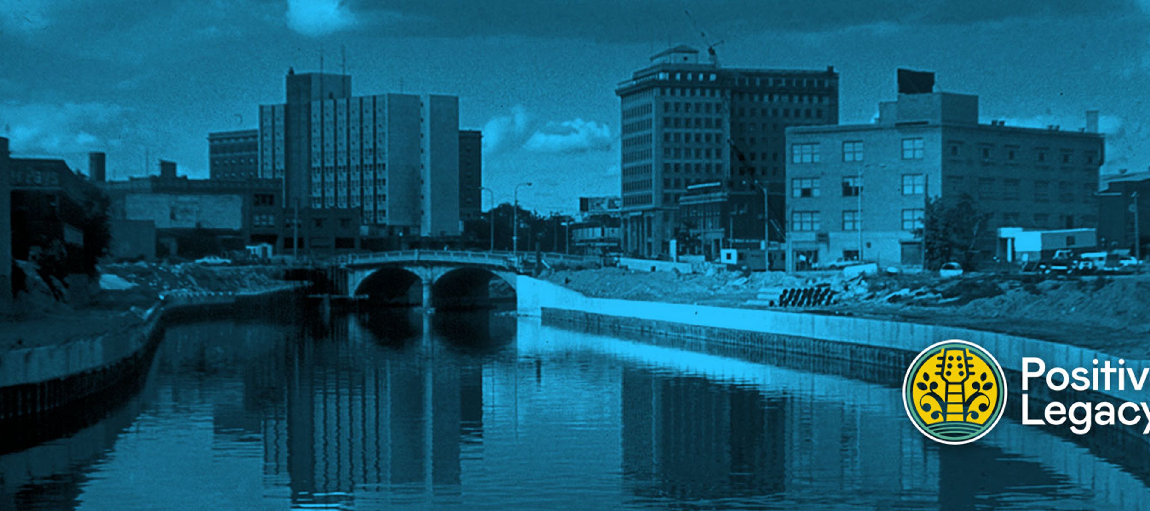 A wide, panoramic landscape photo of a city canal or riverfront, treated with a moody teal-blue tint. The scene features a calm body of water in the foreground reflecting the sky and buildings. A concrete arched bridge crosses the water in the mid-ground. Along the banks, there are several multi-story urban buildings, some appearing older and industrial, set against a sky with soft, scattered clouds.