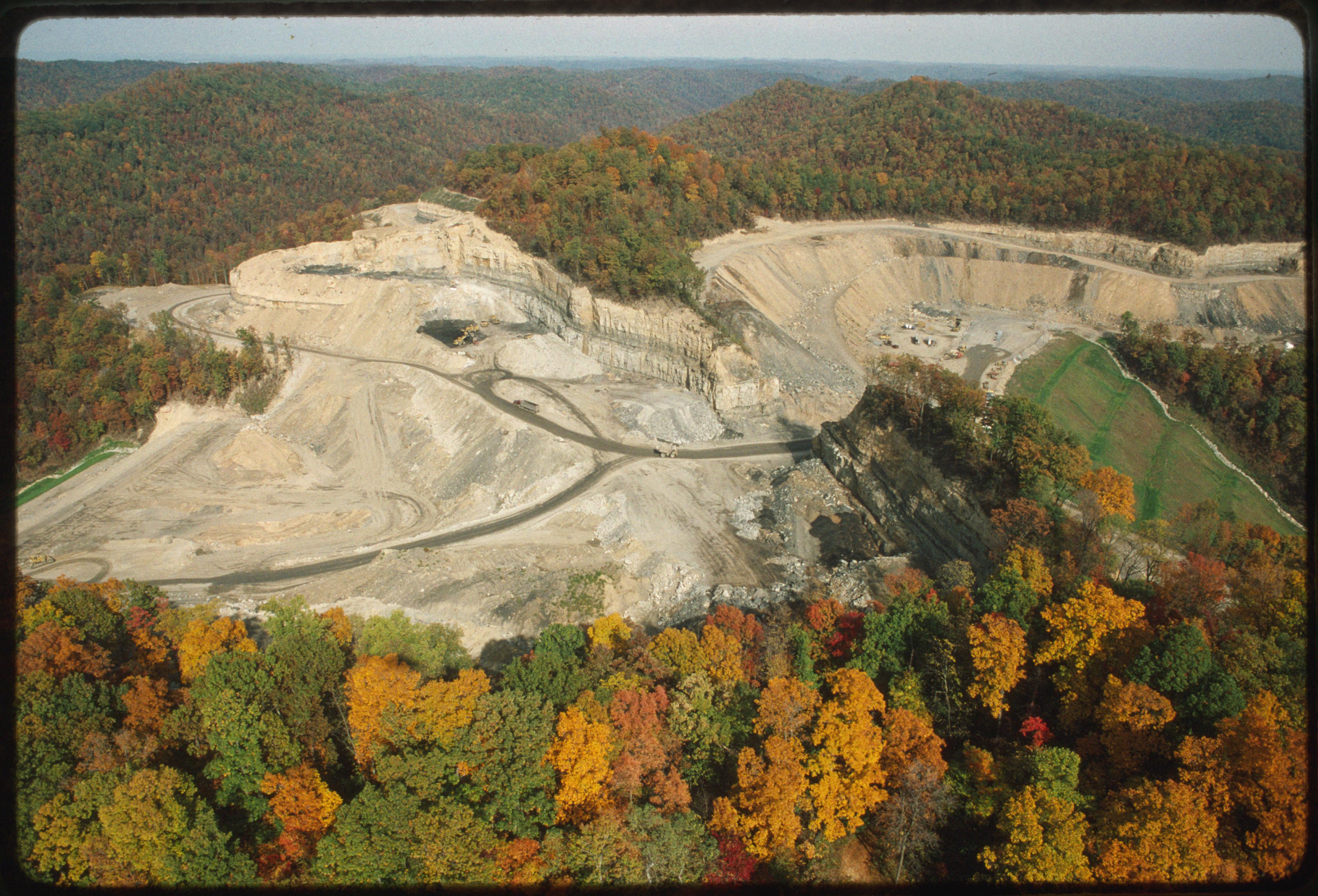 An aerial, high-angle photograph shows an active strip mining site carved into a dense, forested mountain landscape during autumn. The central portion of the image reveals large, exposed layers of rock and soil where the mountainside has been excavated, creating flat terraces and steep, tiered walls. Several haul trucks are visible on dirt paths navigating the mining area. The surrounding terrain is covered in a lush forest displaying vibrant fall colors of green, yellow, orange, and red, providing a stark contrast to the barren, earthy tones of the industrial excavation site.