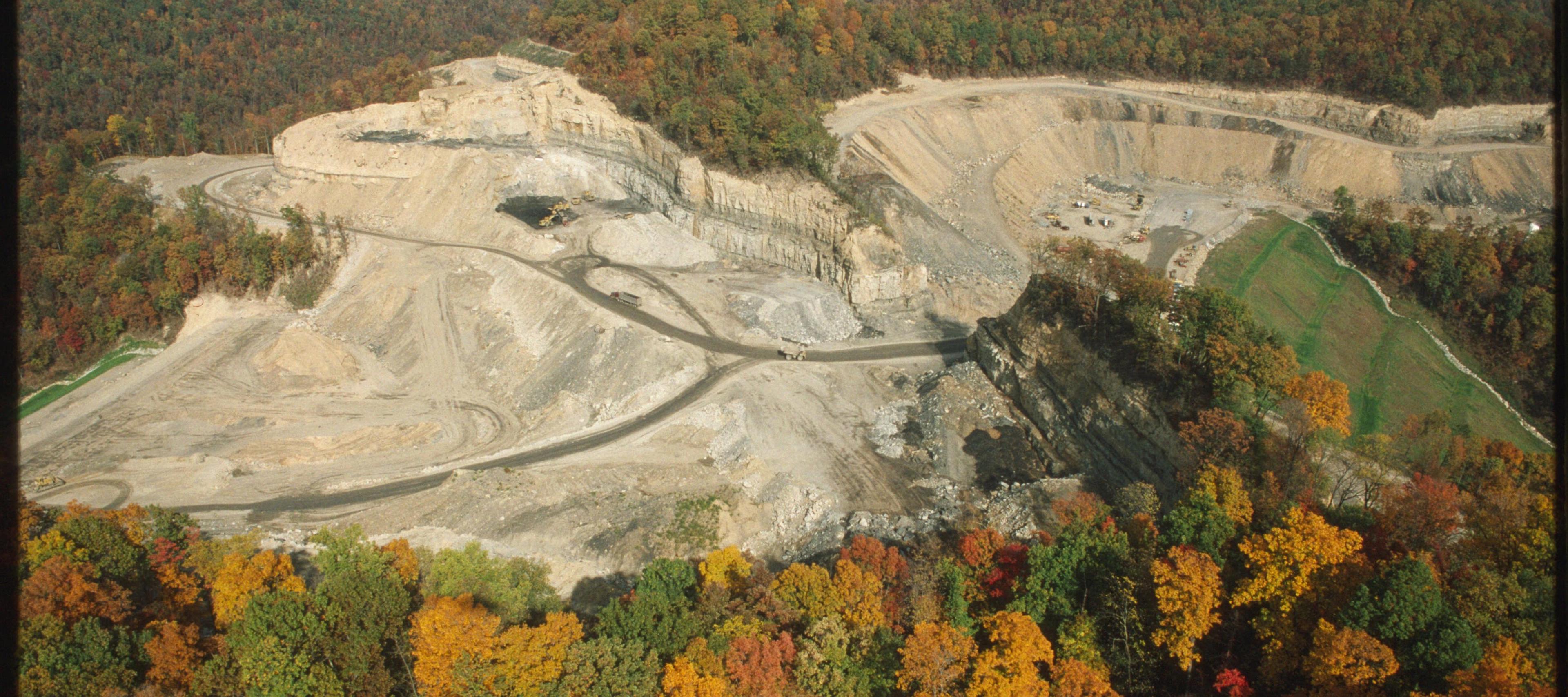 An aerial, high-angle photograph shows an active strip mining site carved into a dense, forested mountain landscape during autumn. The central portion of the image reveals large, exposed layers of rock and soil where the mountainside has been excavated, creating flat terraces and steep, tiered walls. Several haul trucks are visible on dirt paths navigating the mining area. The surrounding terrain is covered in a lush forest displaying vibrant fall colors of green, yellow, orange, and red, providing a stark contrast to the barren, earthy tones of the industrial excavation site.