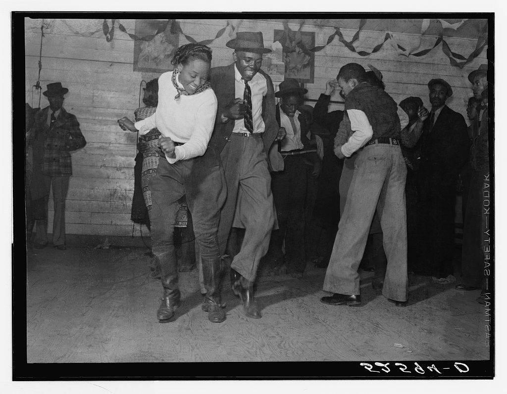 A black and white photograph by Russell Lee titled "Jitterbugging in a juke joint," taken in 1939 in Clarksdale, Mississippi.  Central Figures: The focus is on a Black man and woman dancing energetically in the center of a crowded room. The woman, on the left, wears a light-colored sweater, dark breeches, and tall dark boots with a patterned scarf around her neck. The man, on the right, wears a dark suit jacket, a necktie, and a fedora. Both have joyous expressions as they kick their legs and swing their arms in rhythm.